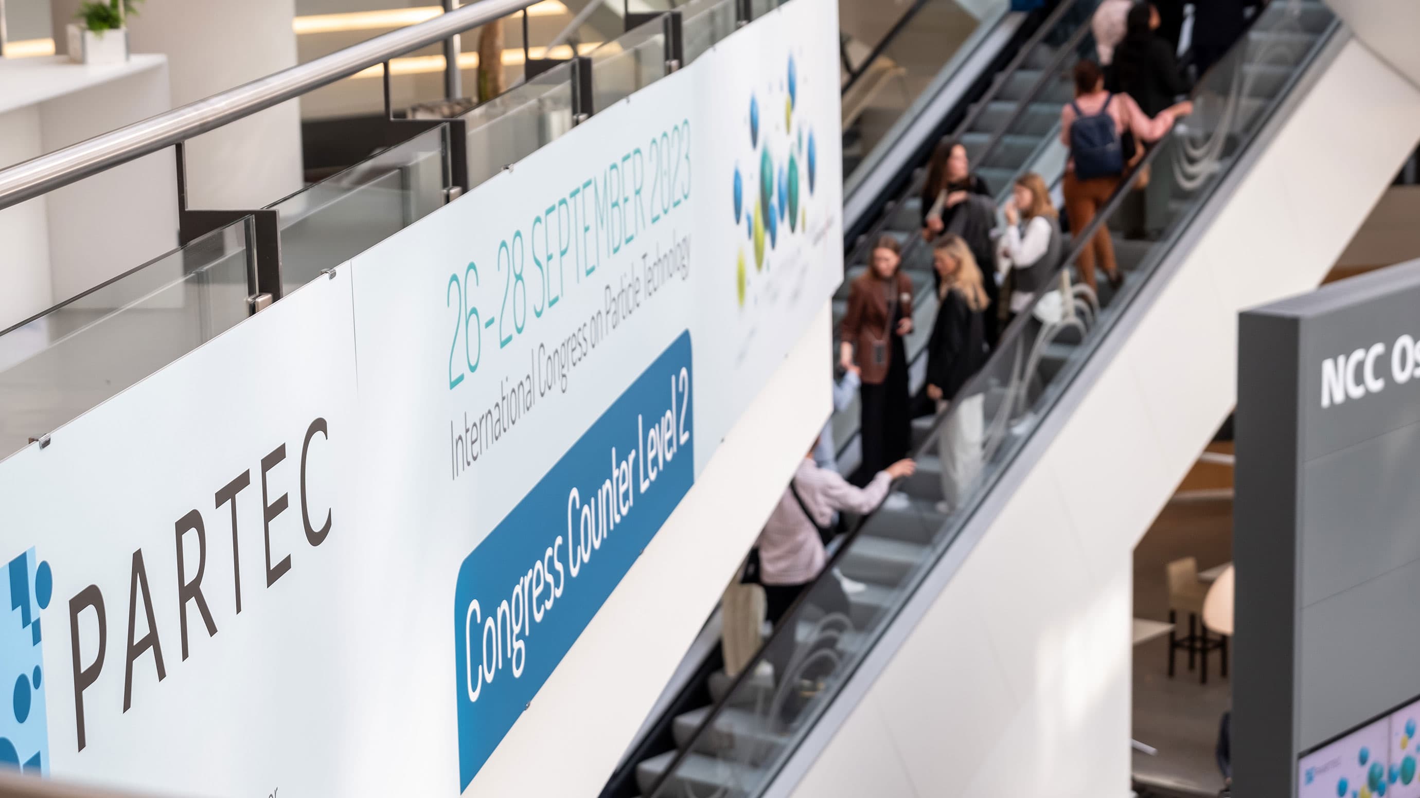 PARTEC banner with an escalator full of people in the background